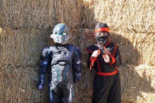 Two children in costumes are standing next to each other in front of a hay bale.