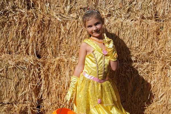 A little girl in a yellow dress and gloves is standing in front of a hay bale.