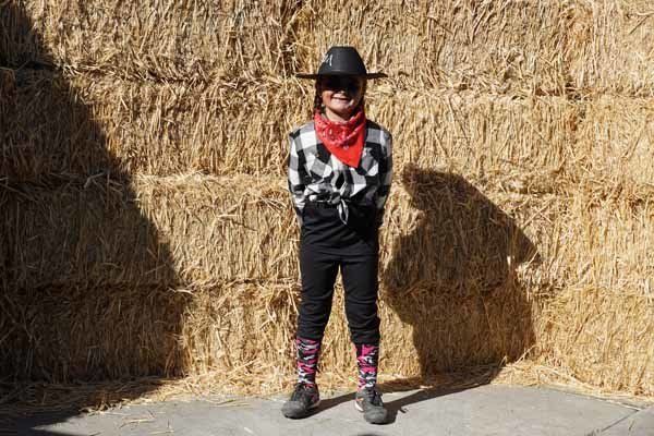 A young boy dressed as a cowboy is standing in front of a hay bale.