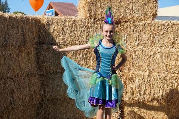 A girl in a peacock costume is standing in front of hay bales.