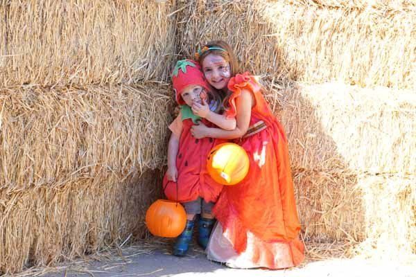 Two little girls in tomato costumes are hugging each other in front of a hay bale.