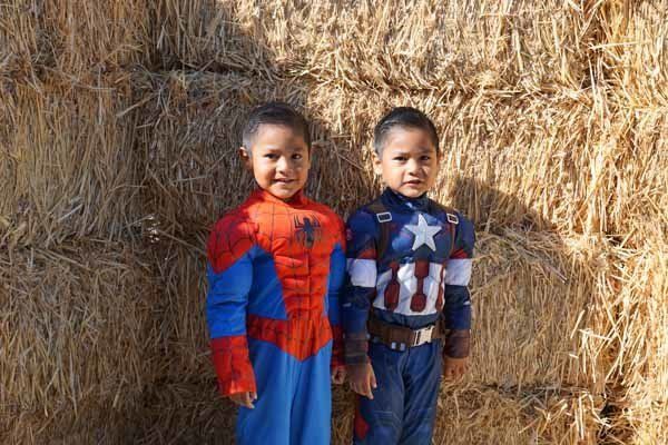 Two young boys in superhero costumes are standing next to each other in front of a hay bale.