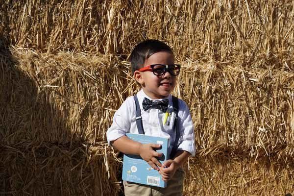 A little boy wearing sunglasses and suspenders is standing in front of a hay bale.
