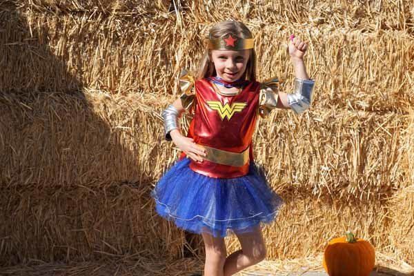 A little girl in a wonder woman costume is standing in front of a hay bale.