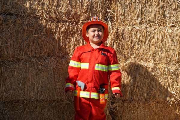 A young boy in a fireman costume is standing in front of a hay bale.