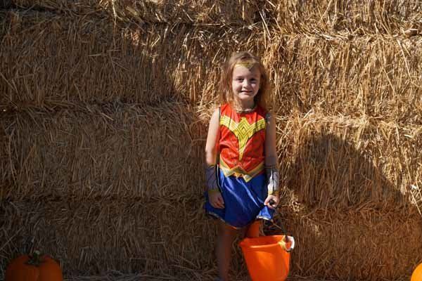 A little girl in a wonder woman costume is standing in front of a hay bale.