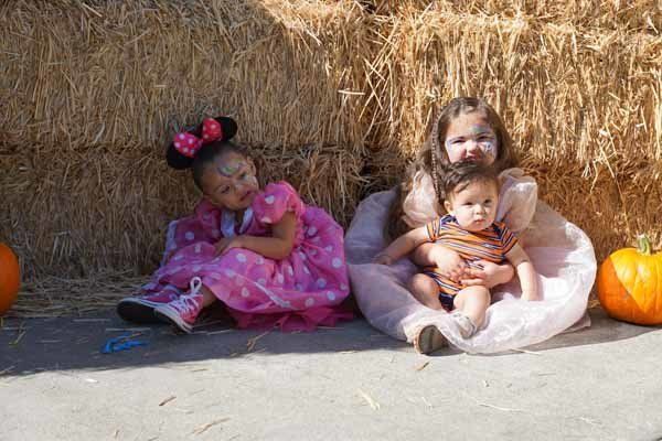 Two little girls and a baby are sitting in front of a hay bale.