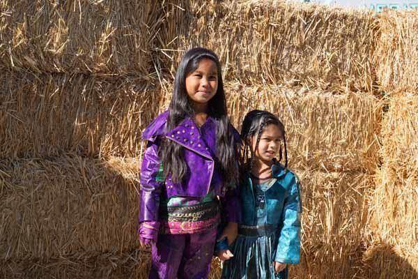 Two young girls are standing next to each other in front of a pile of hay.