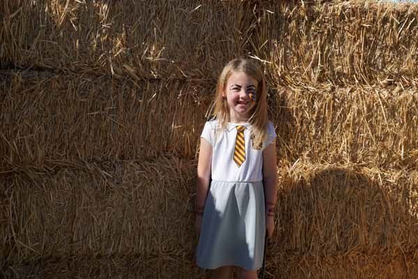 A little girl in a dress and tie is standing in front of a pile of hay.