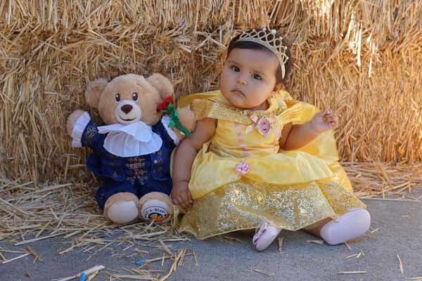 A baby dressed as belle is sitting next to a teddy bear