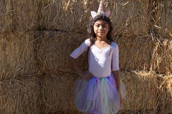 A little girl dressed as a unicorn is standing in front of a hay bale.