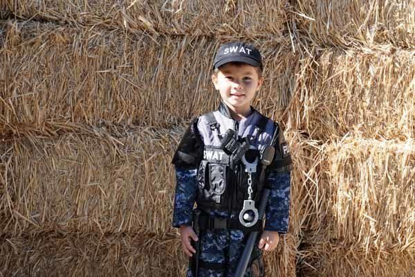 A young boy in a swat costume is standing in front of a bale of hay.