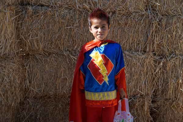 A young boy in a superhero costume is standing in front of a hay bale.