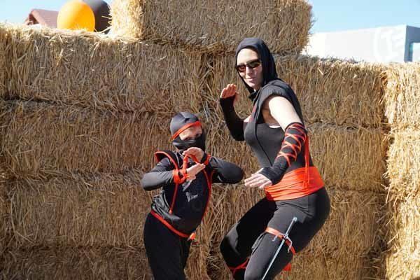 A woman and a child dressed as ninjas are jumping in front of hay bales.