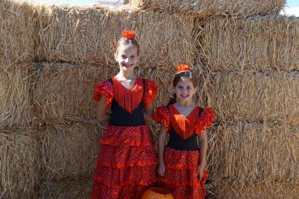 Two little girls in red dresses are standing next to each other in front of hay bales.