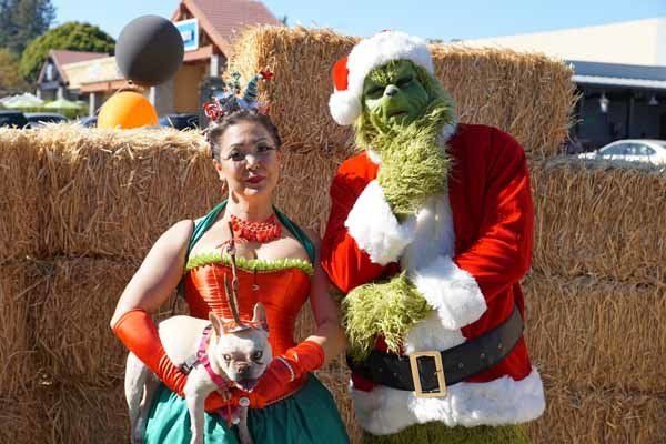 A woman is standing next to a man dressed as grinch holding a dog.
