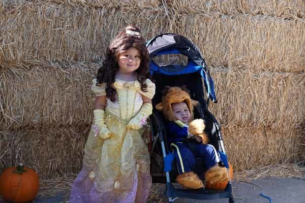 A little girl dressed as belle is standing next to a baby in a stroller.