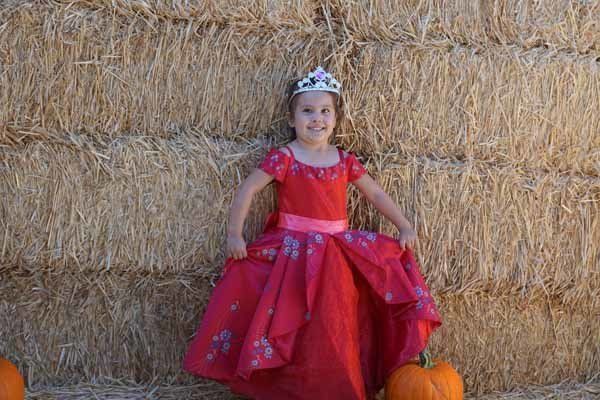 A little girl in a red dress and tiara is standing in front of a hay bale.
