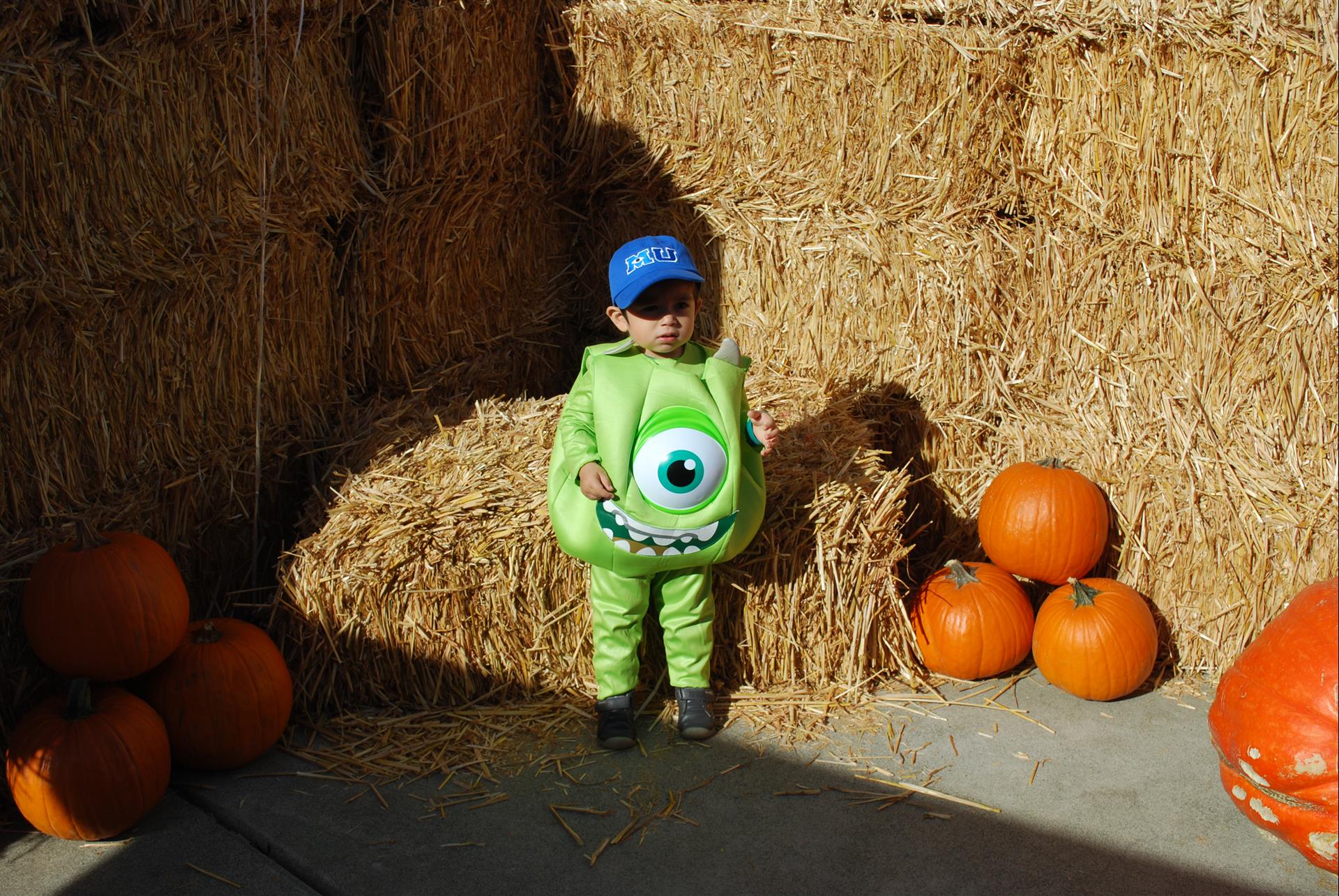 A little boy in a mike wazowski costume is standing next to hay bales and pumpkins.