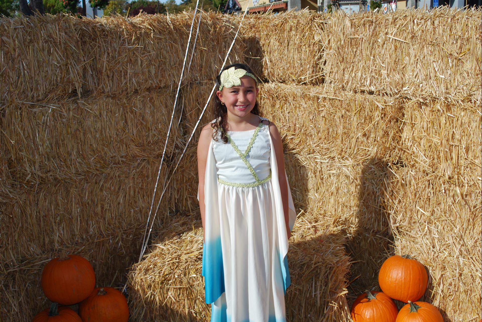 A little girl in a dress is standing in front of hay bales and pumpkins.