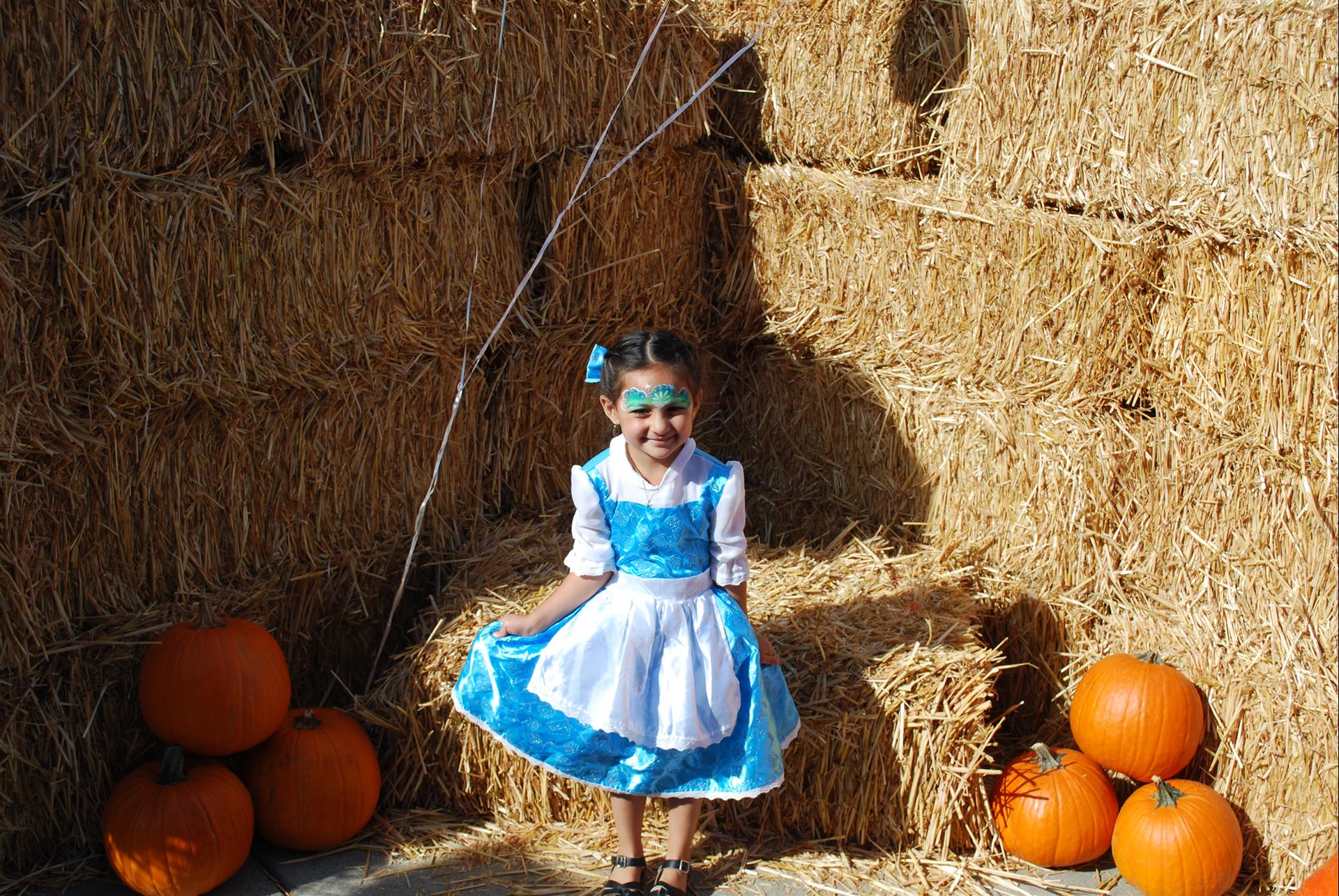 A little girl in a blue dress is standing in front of hay bales and pumpkins.