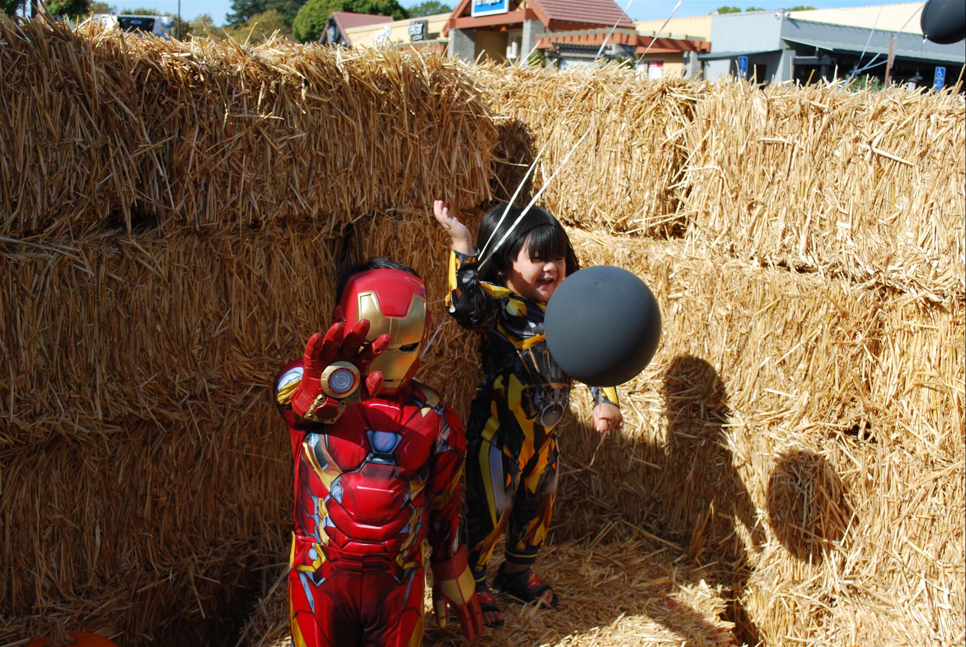 Two children in iron man costumes are playing with balloons in a hay bale maze.