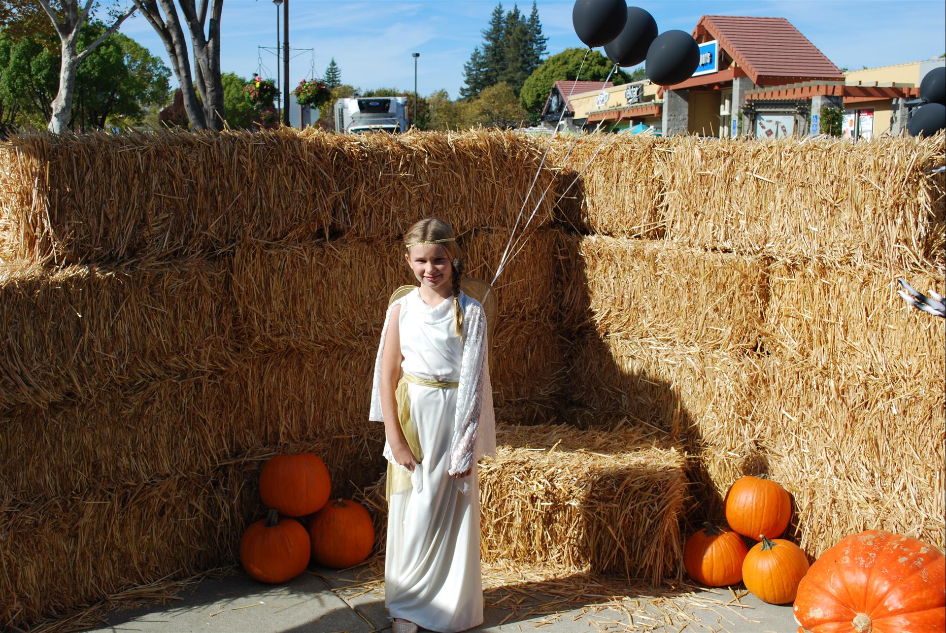 A girl in a white dress stands in front of hay bales and pumpkins
