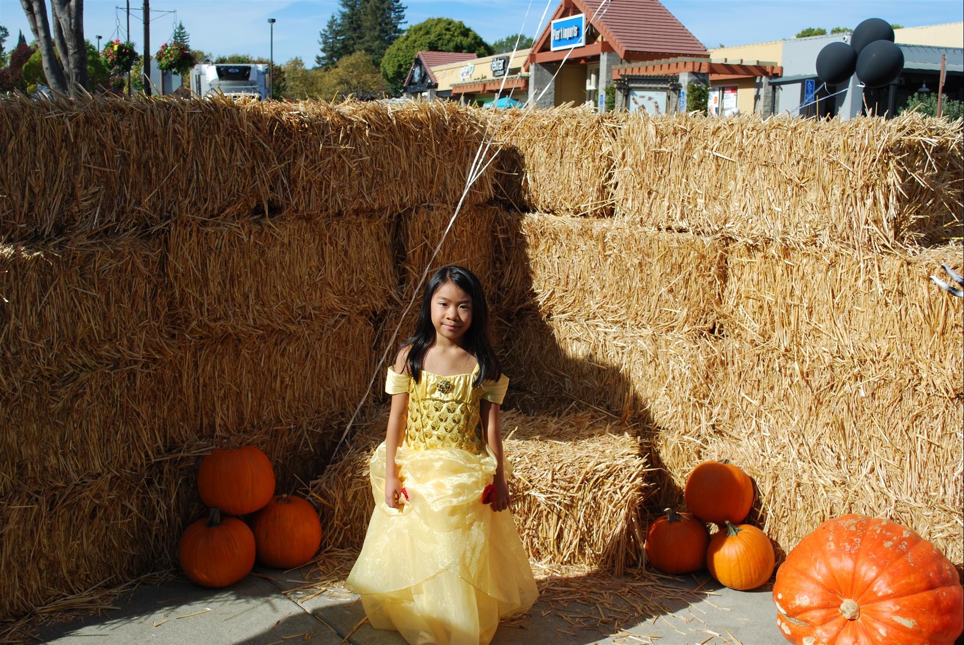 A little girl in a yellow dress is standing in front of hay bales and pumpkins.