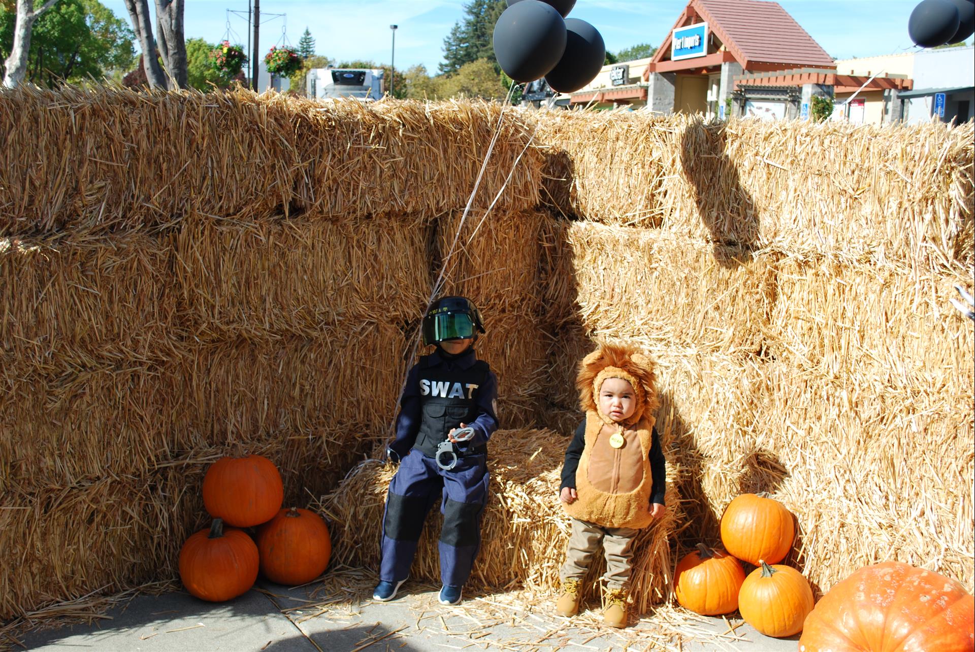 Two children in swat costumes are sitting on hay bales surrounded by pumpkins
