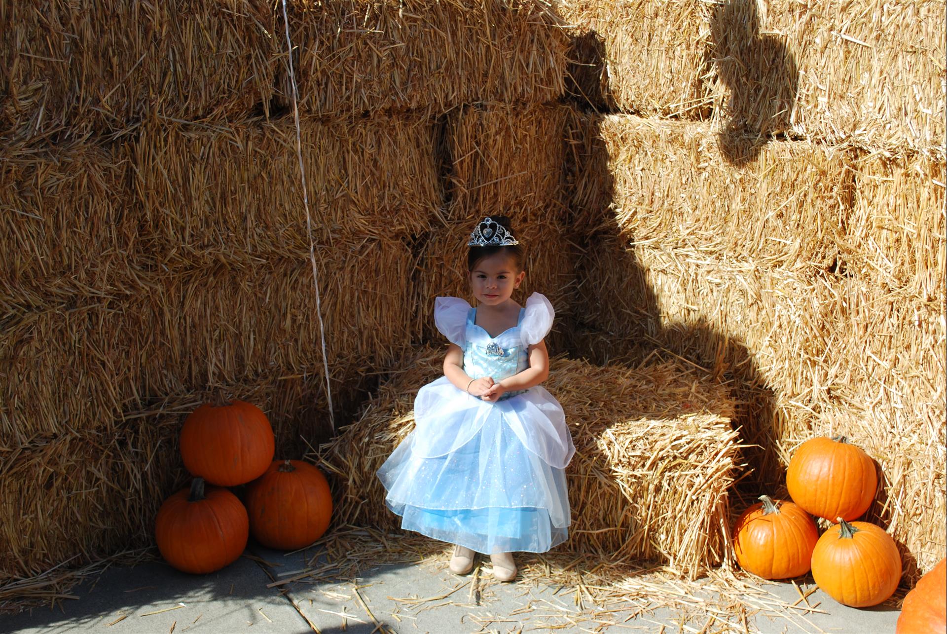 A little girl dressed as cinderella is sitting on a bale of hay surrounded by pumpkins.