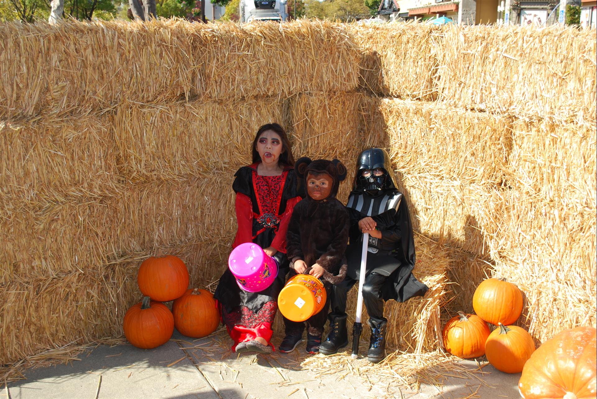 Three children dressed in halloween costumes are sitting on hay bales surrounded by pumpkins.
