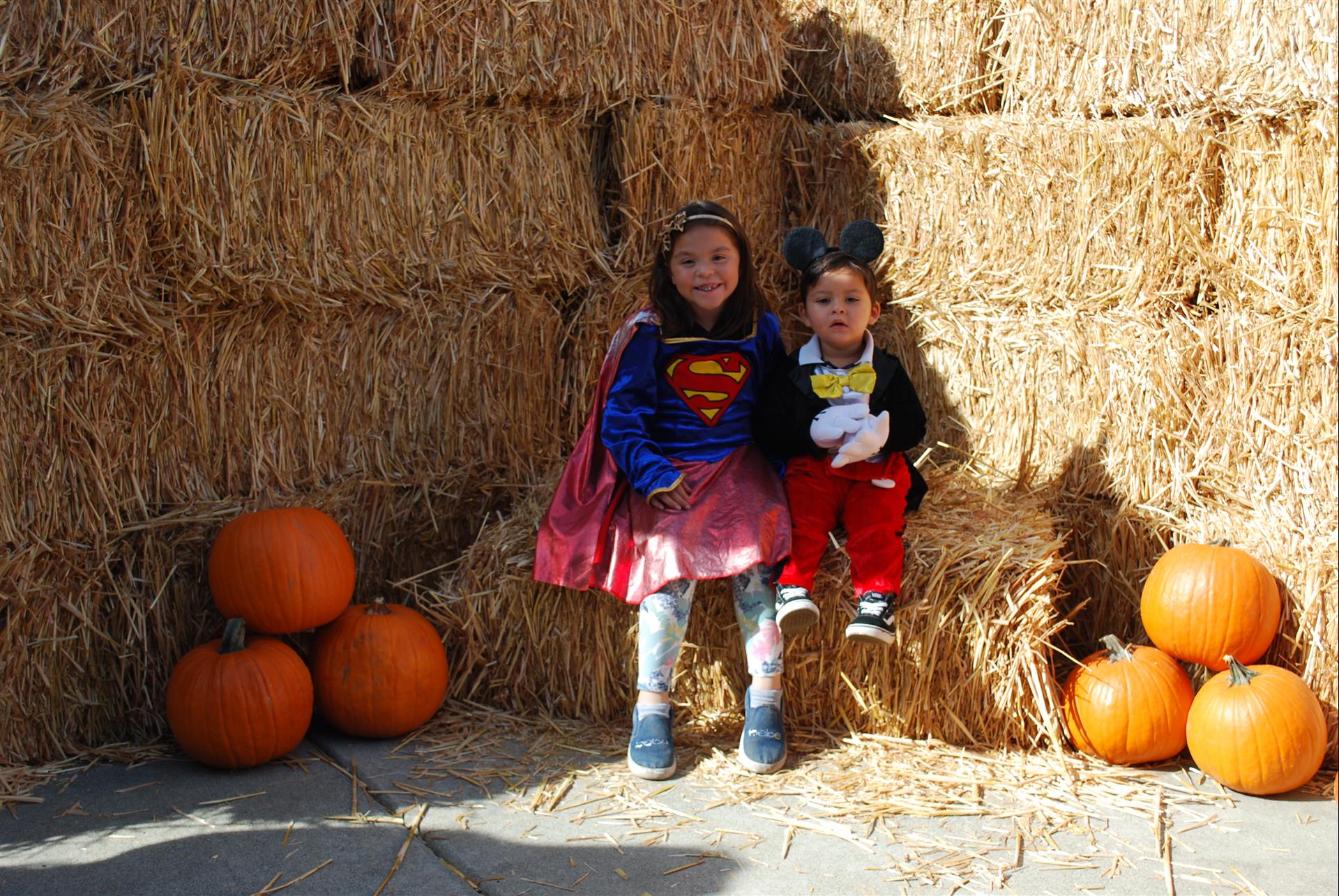 Two children in costumes are sitting on bales of hay surrounded by pumpkins.