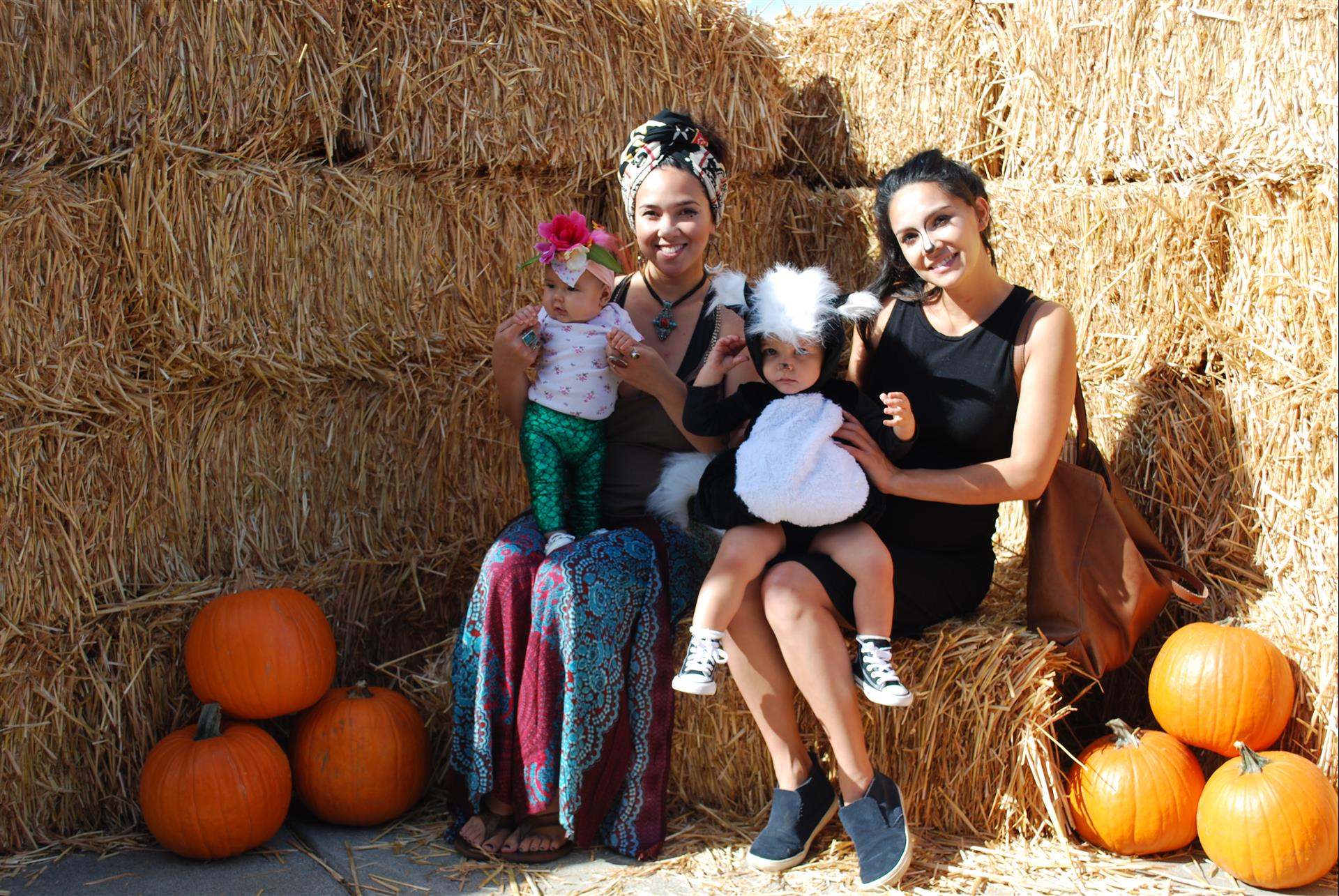 Two women and a baby are sitting on a bale of hay surrounded by pumpkins.