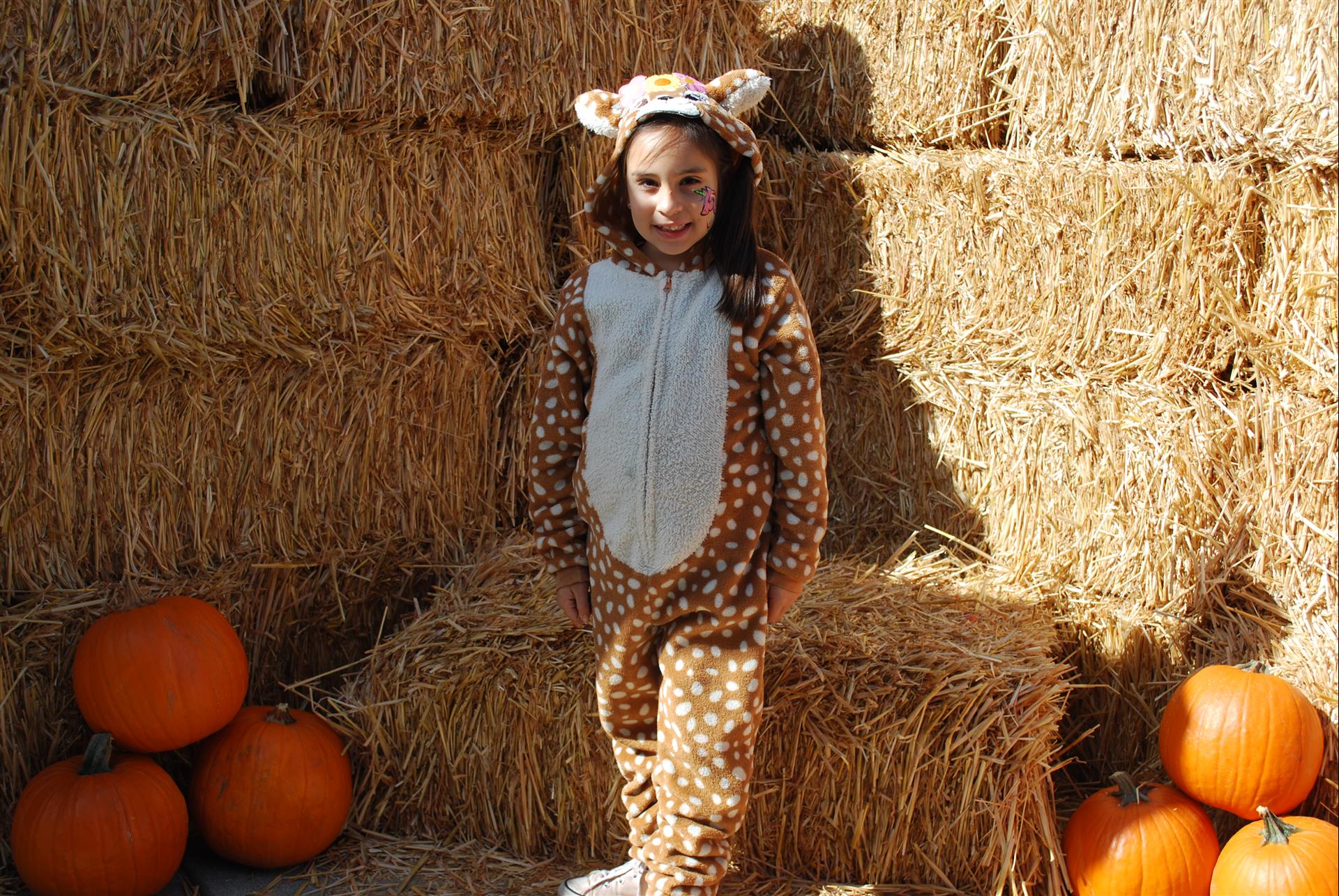 A little girl in a deer costume is sitting on a bale of hay surrounded by pumpkins.