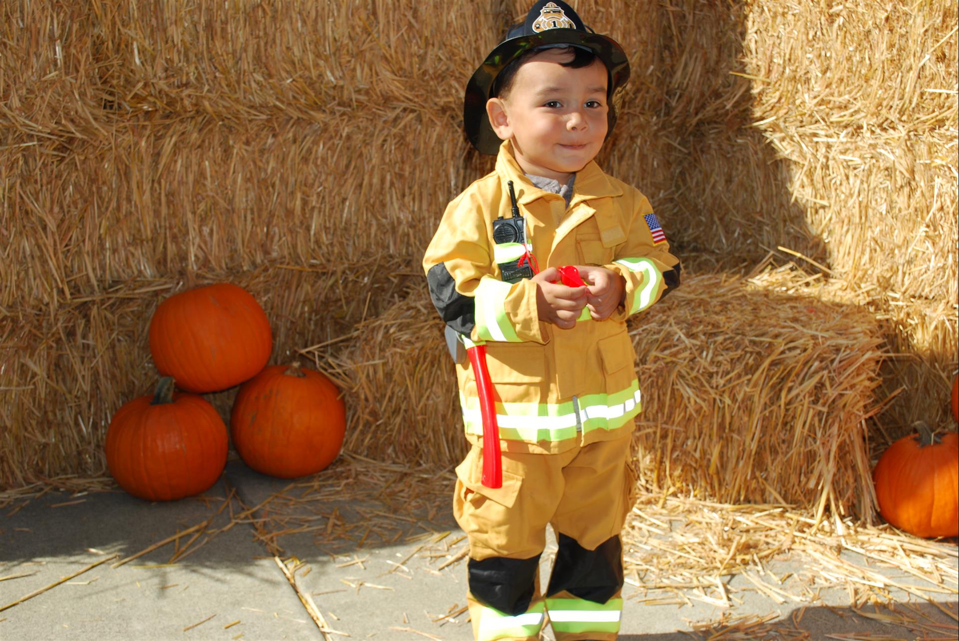 A little boy in a fireman costume is standing in front of hay bales and pumpkins