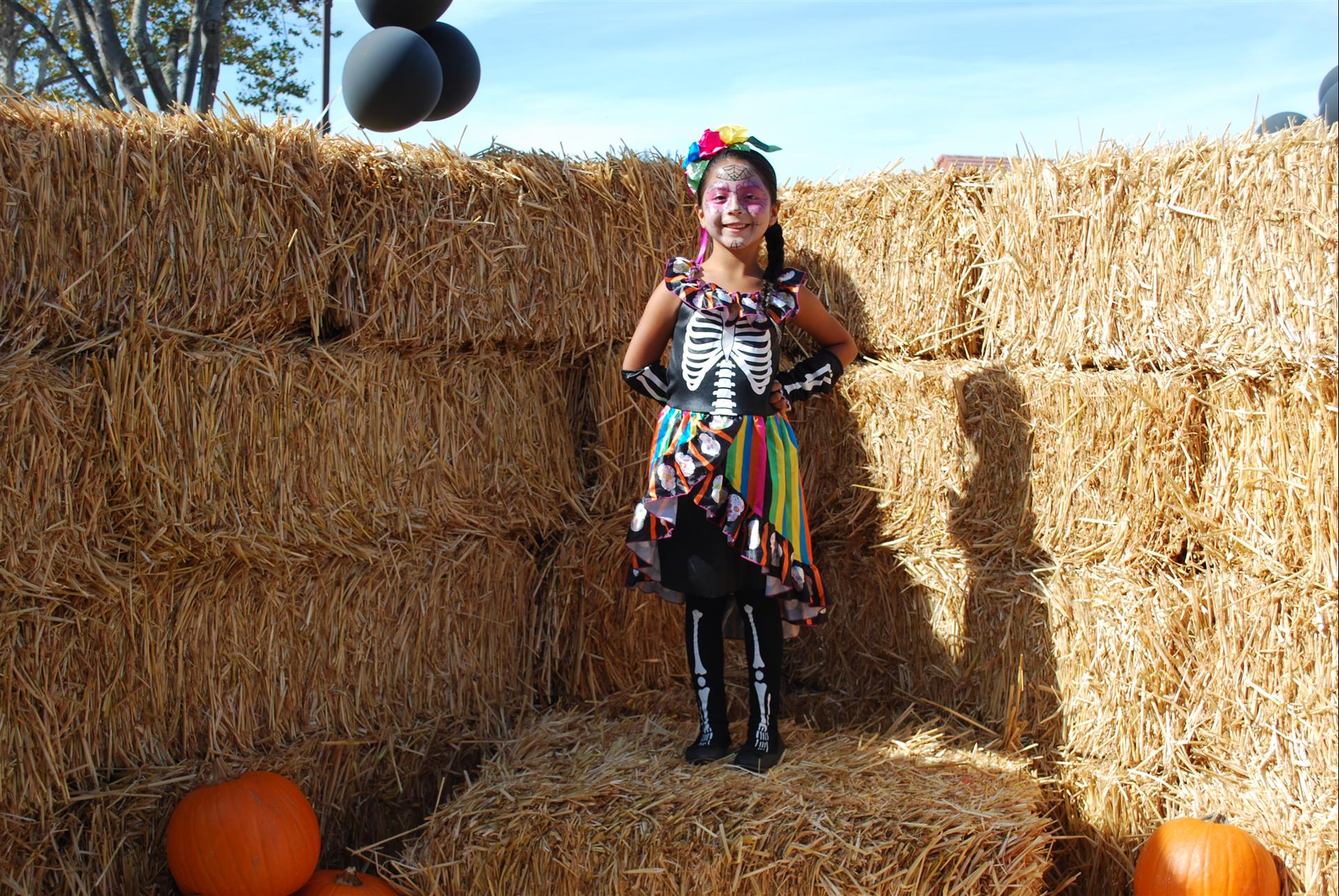 A little girl in a skeleton costume is standing next to hay bales and pumpkins.