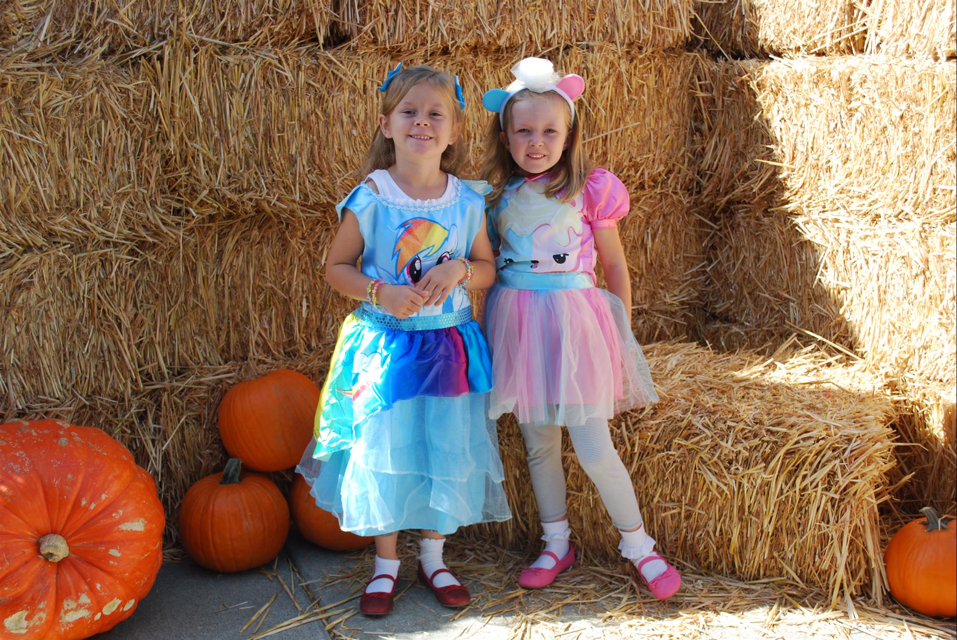 Two little girls are posing for a picture in front of hay bales and pumpkins.