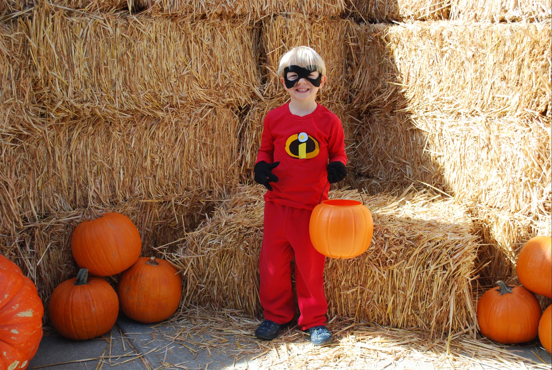 A boy in a superhero costume is holding a pumpkin in front of hay bales.