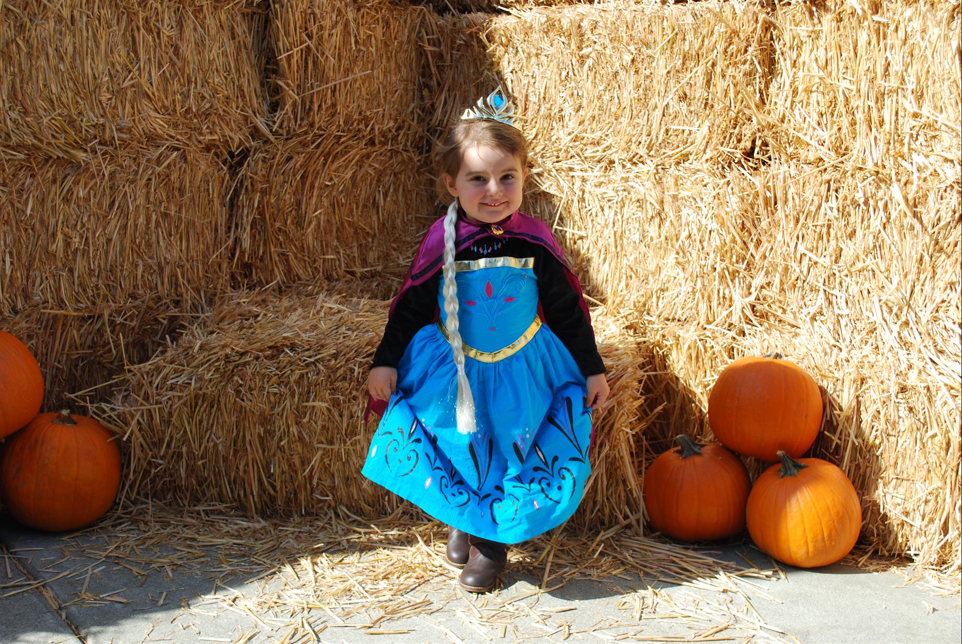 A little girl dressed as elsa from frozen is standing in front of hay bales and pumpkins.