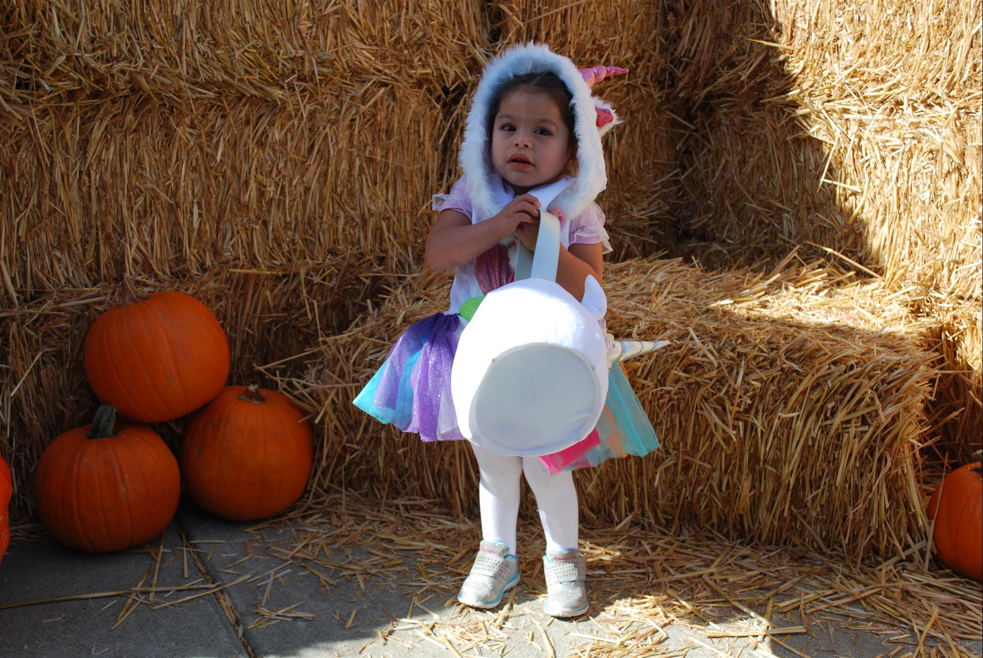 A little girl in a unicorn costume is standing next to hay bales and pumpkins.