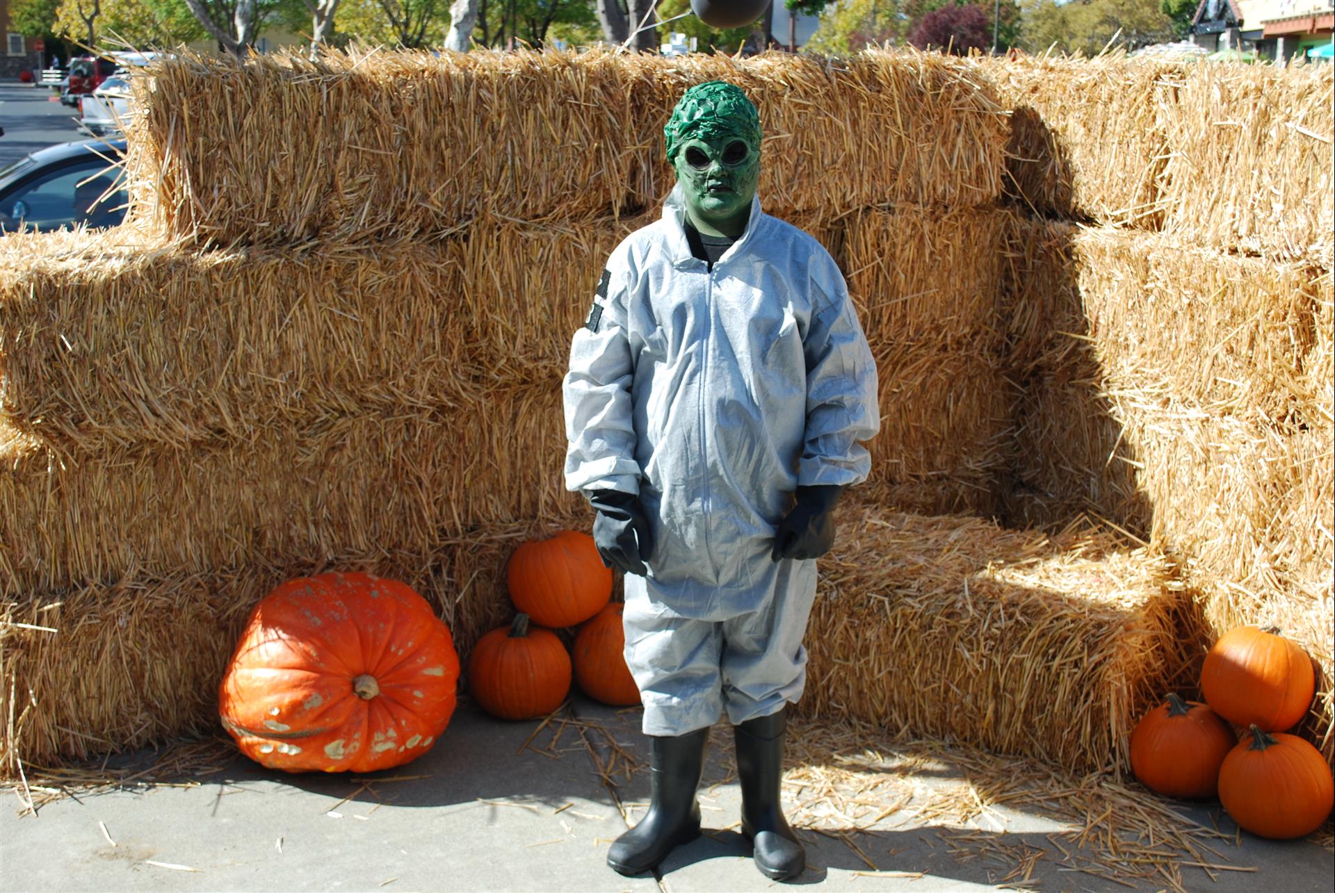 A person in a green mask is standing in front of hay bales and pumpkins.