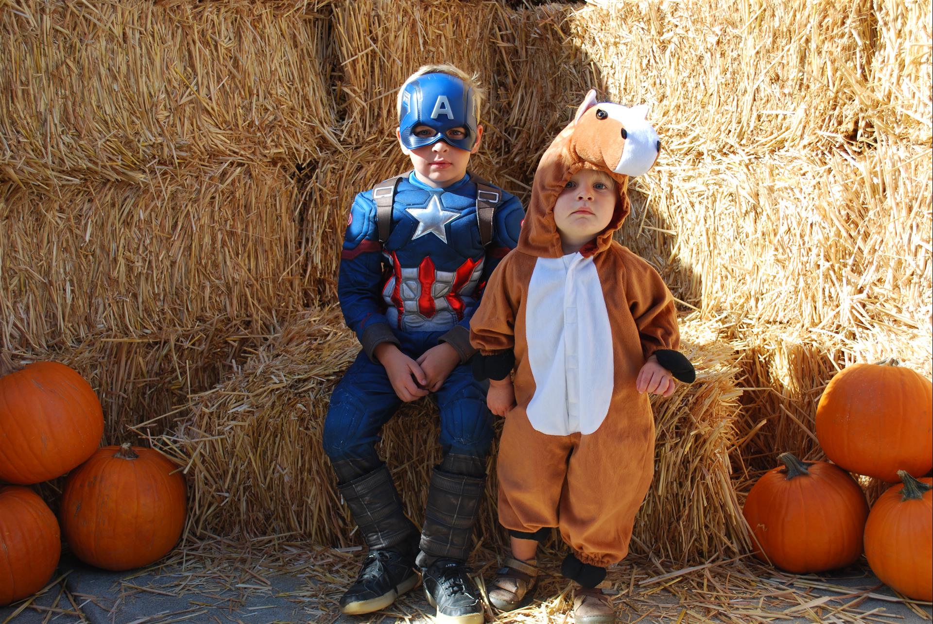 A boy dressed as captain america and a boy dressed as a horse are sitting on a bale of hay.