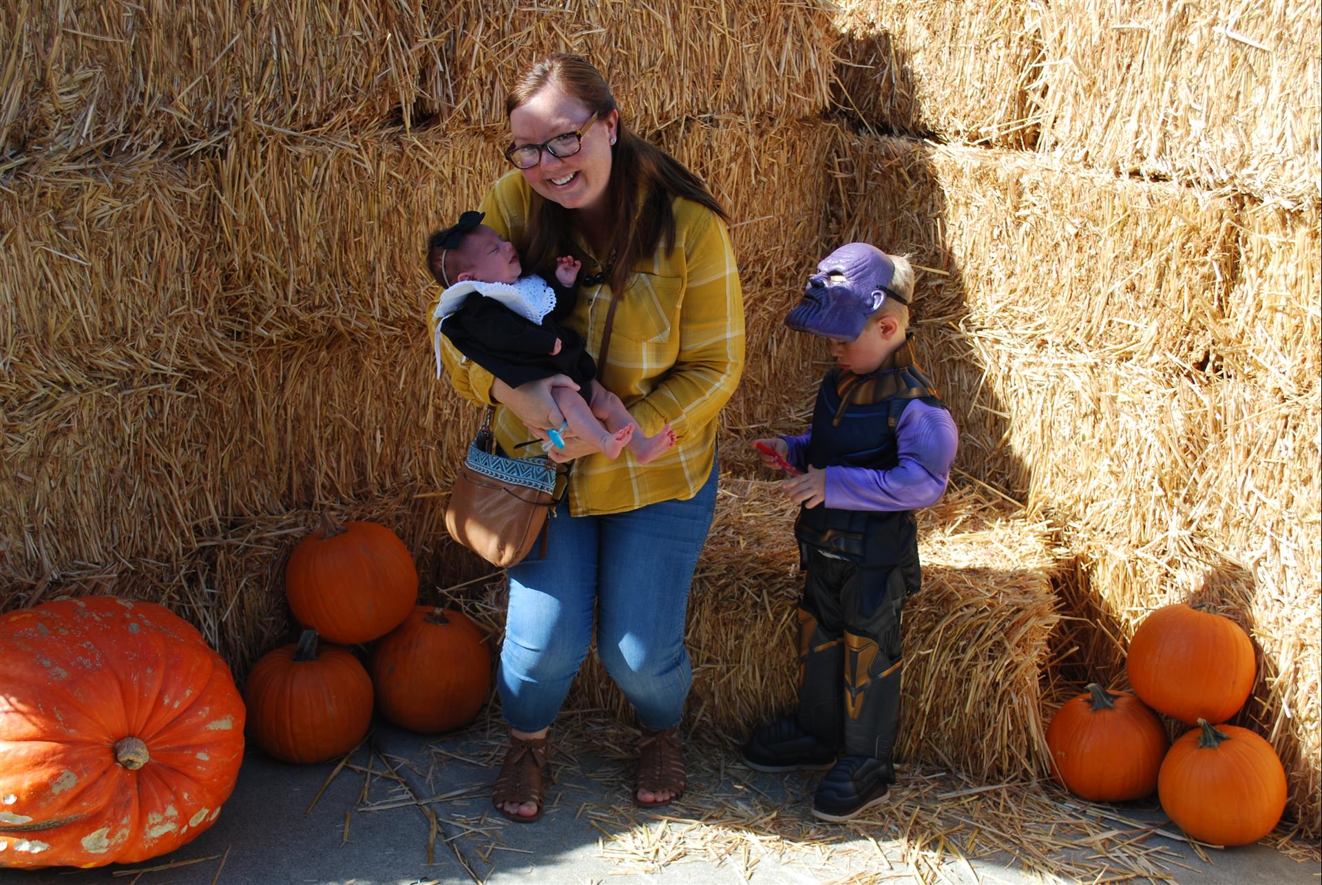 A woman is holding a baby and two children are standing next to hay bales.