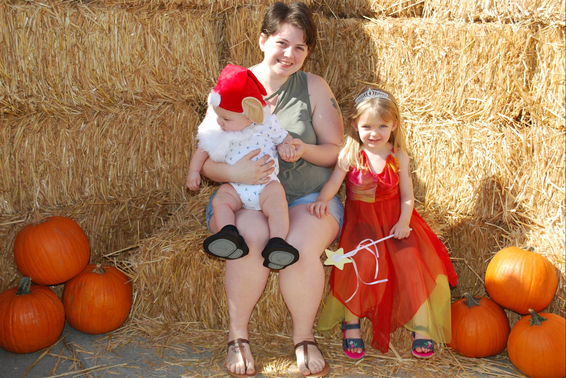 A woman is holding a baby and two little girls are sitting on hay bales
