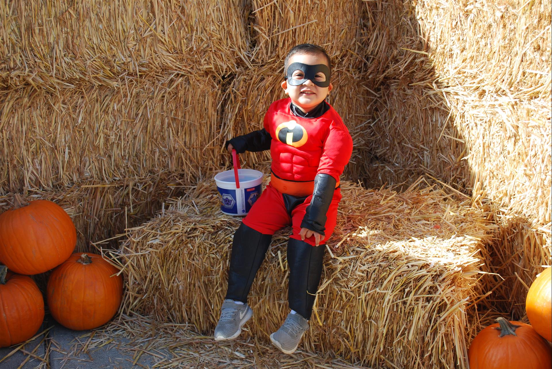 A little boy in a superhero costume is sitting on a bale of hay.