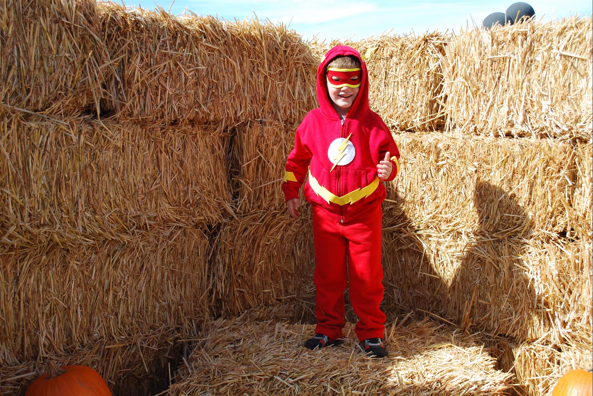 A young boy in a flash costume is standing in a pile of hay.