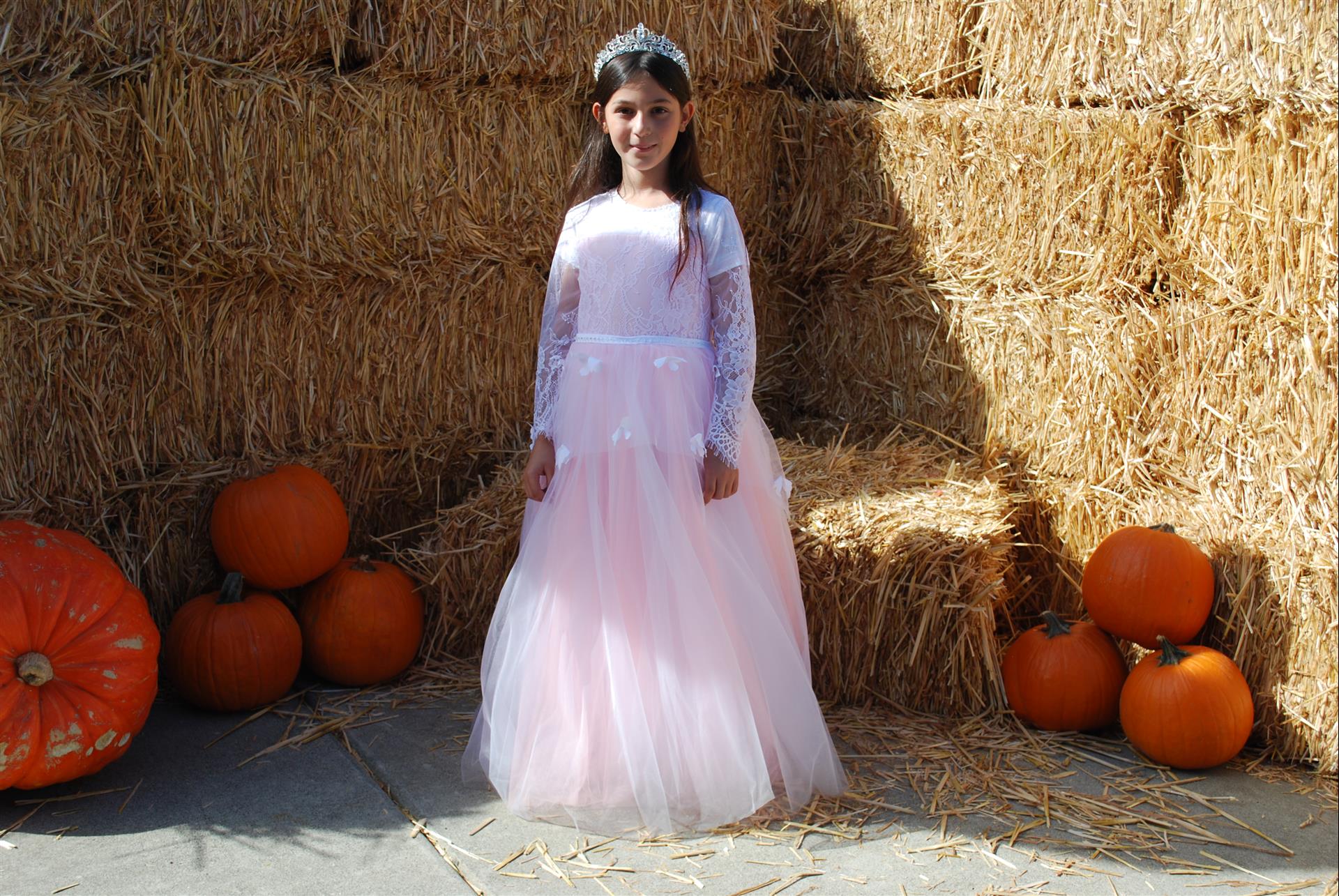 A little girl in a pink dress is standing in front of hay bales and pumpkins.