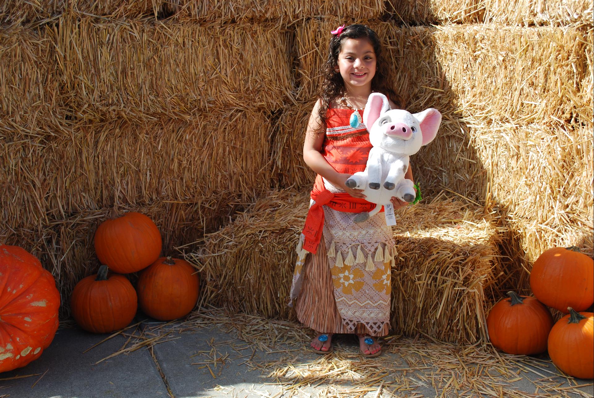 A little girl is sitting on a bale of hay holding a stuffed animal.