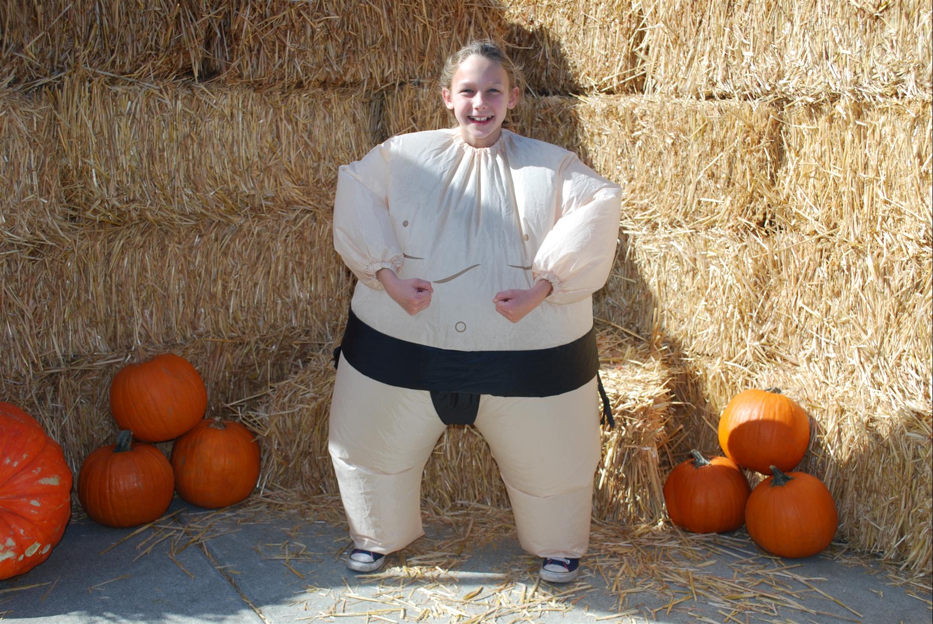A woman in an inflatable sumo wrestler costume is standing in front of hay bales and pumpkins.