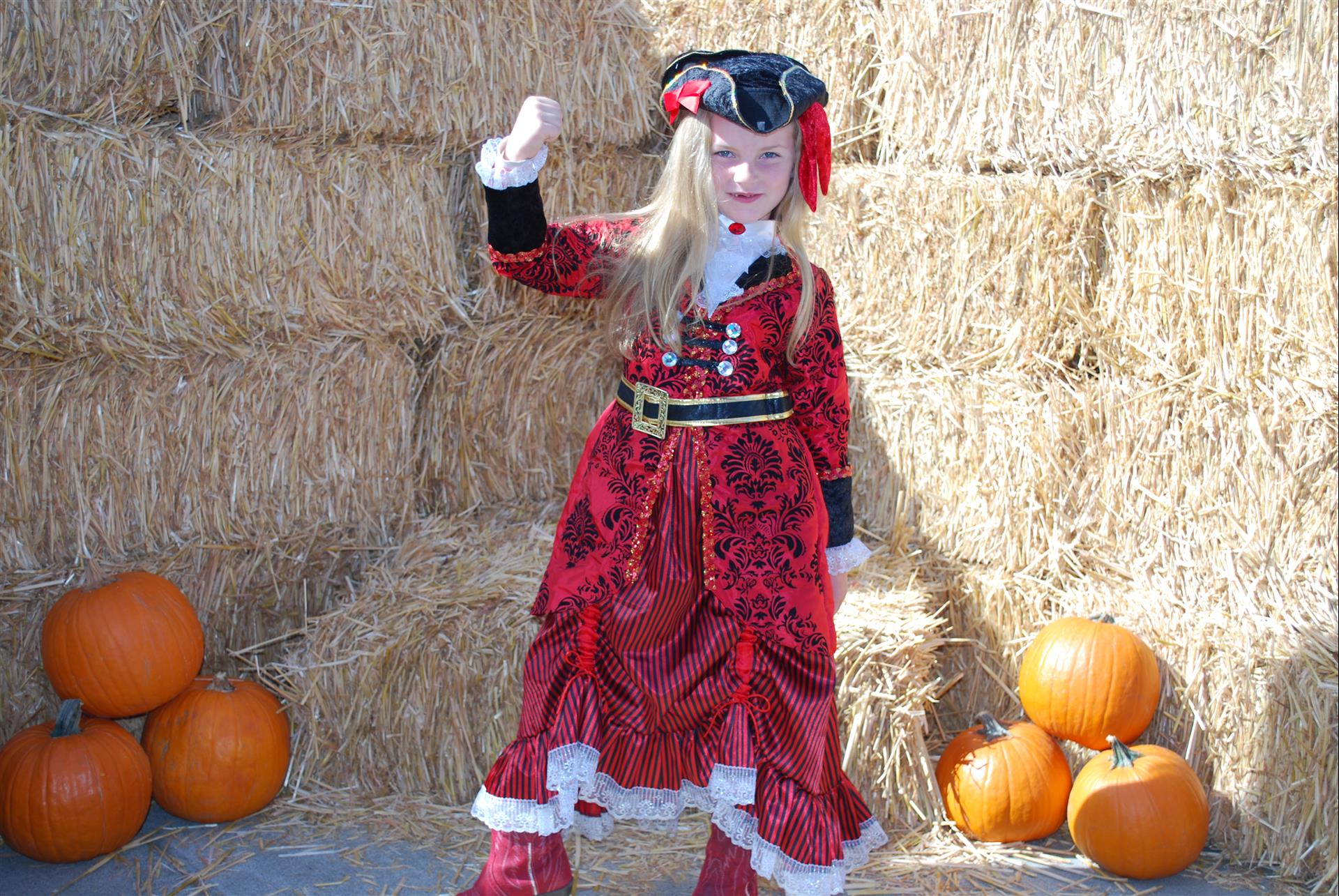 A little girl in a pirate costume is standing in front of hay bales and pumpkins.